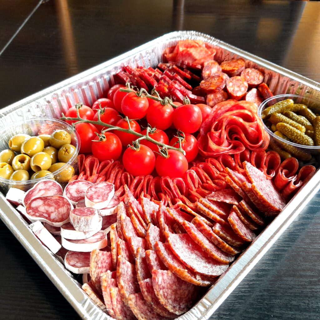 Na stole je podnos s masem a zeleninou: 5 druhů salámu, olivy, Cherry rajčátka a okurčičky.